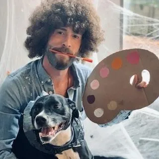 Brandon George in a Bob Ross wig, holding a paint palette, with a grinning black-and-white dog under one arm